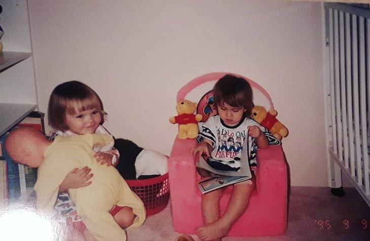 A picture of three-year-old me sitting in a fuzzy pink chair reading a book while a friend plays with a doll next to me.