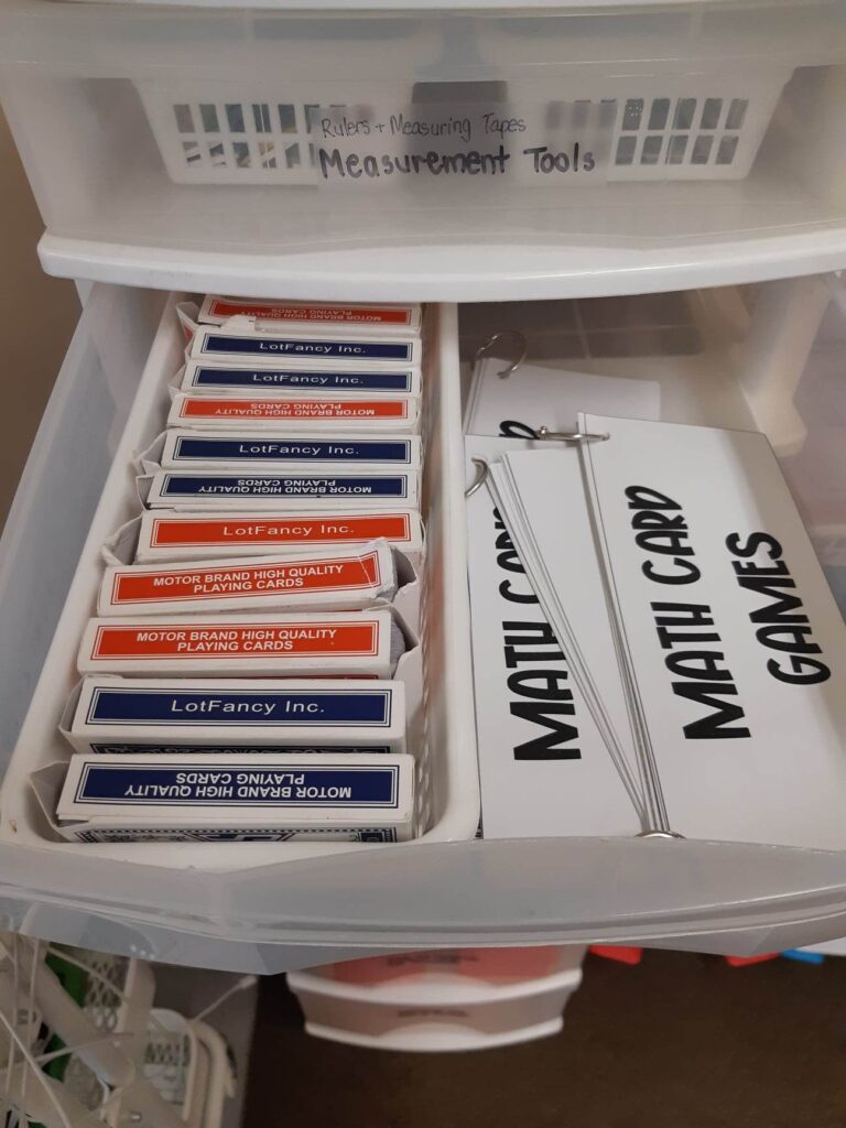 A set of plastic drawers. One drawer is open. On the left side, there is a plastic bin with several decks of cards. On the right side, there are cardstock cards on a key ring that read "math card games"